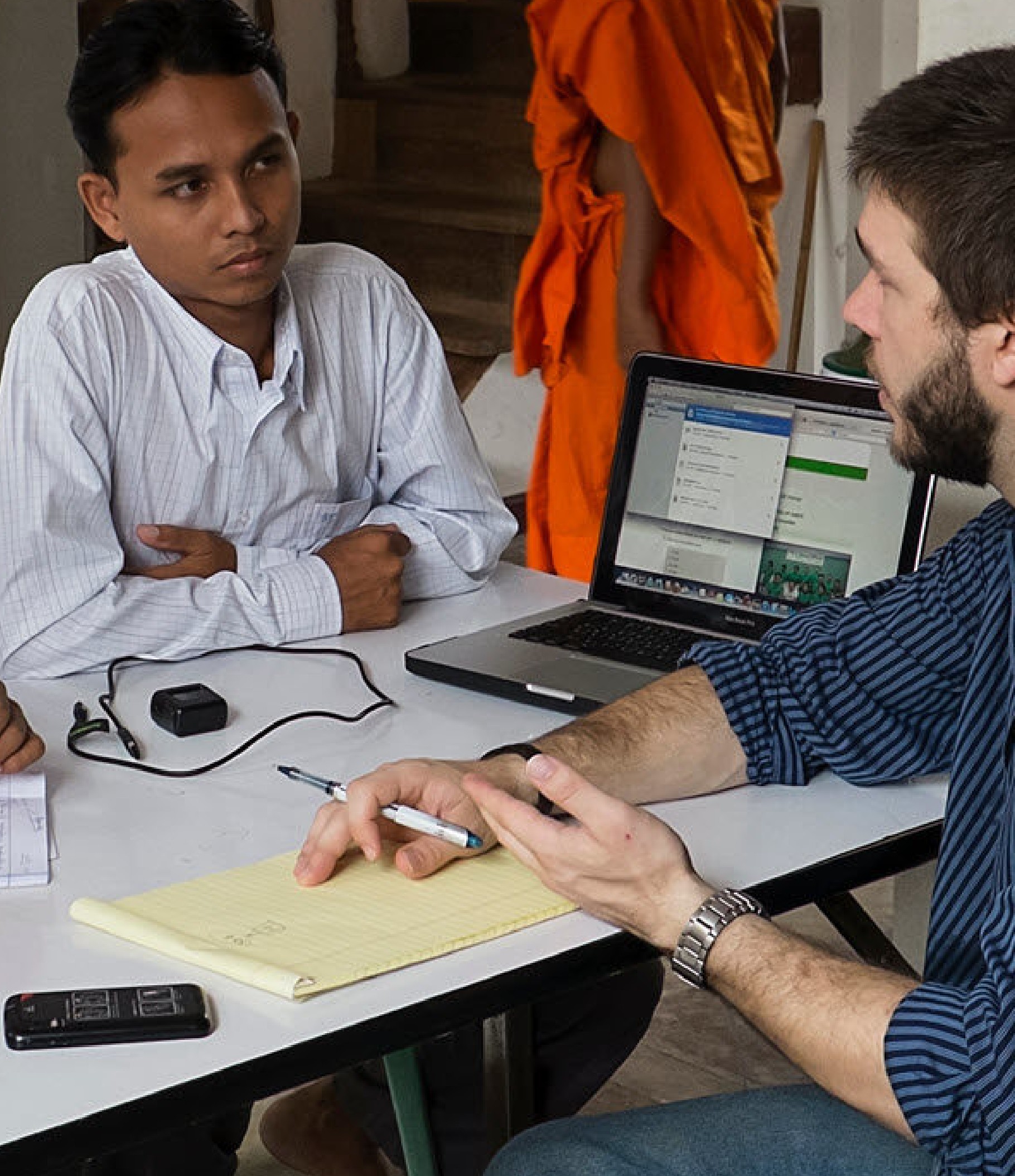 A white man interviews a Burmese man, seated at a desk with laptop and mobile phone